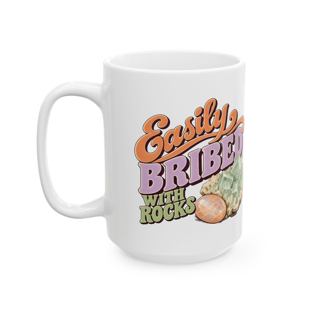 White Easily Bribed with Rocks ceramic mug with retro orange and purple lettering, green crystal artwork and an orange stone — handle-left view.
