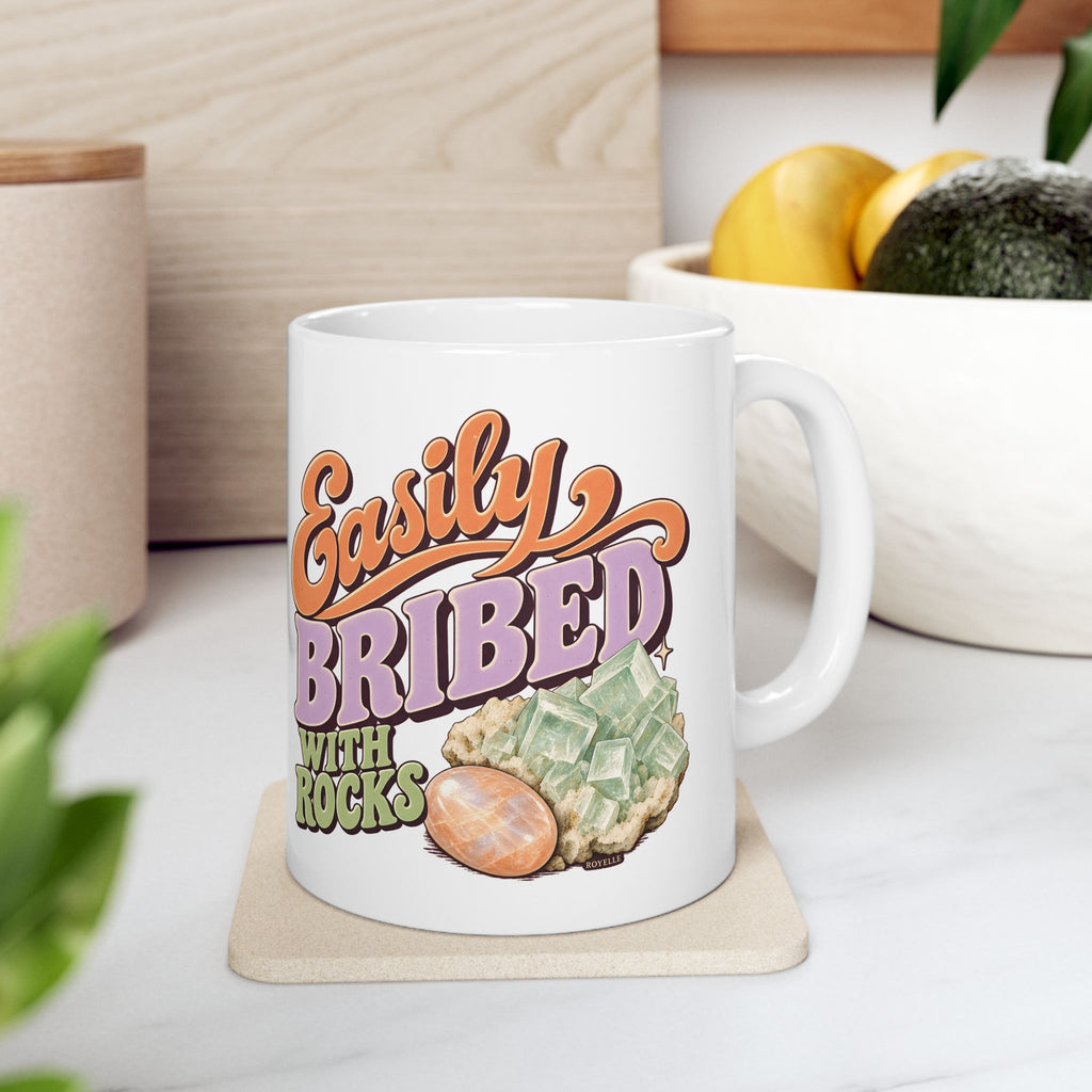 White Easily Bribed with Rocks ceramic mug with retro orange and purple lettering, green crystal artwork and an orange stone — kitchen lifestyle scene; other items shown are props.